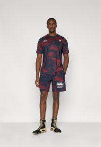 Σετ αθλητικών ρούχων σε μπορντό και ναυτικό μπλε, που περιλαμβάνει μπλούζα με λαιμόκοψη και ταιριαστά σορτς με γραφικό λογότυπο· συνδυάζεται με παπούτσια σε μαύρο και χρυσό χρώμα.