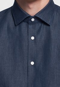 Nahaufnahme einer Person, die ein dunkelblaues, knopfbares Hemd mit spitzem Kragen trägt, an dem drei sichtbare weiße Knöpfe geschlossen sind.