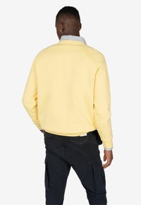 Sudadera color amarillo claro con cuello a rayas, con mangas raglán. Combinada con pantalones cargo oscuros, que muestran detalles de bolsillos y textura.