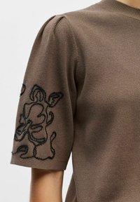 Haut marron en tricot à manches courtes avec détail froncé à l'épaule et broderie florale abstraite noire sur la manche.