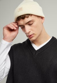 Black knitted sleeveless sweater over a white long-sleeve shirt, paired with a ribbed cream beanie featuring a small logo patch.
