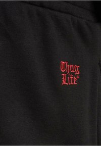 Svart stoff med en myk tekstur, med røde broderte bokstaver "Thug Life" foran, som fremhever designet.