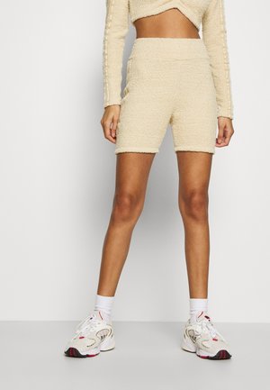Femme portant un short de motard beige texturé, un haut assorti à manches longues raccourci, des chaussettes blanches et des baskets blanches de sport, debout devant un fond uni.