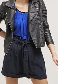 Veste en cuir noir avec quincaillerie argentée, haut sans manches bleu avec plis, et shorts taille haute bleu marine avec ceinture à nouer et manches retroussées.