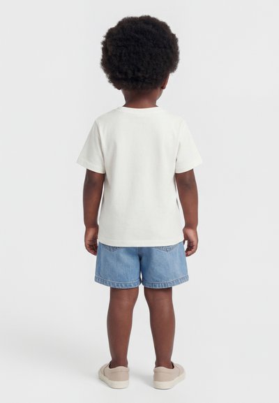 Kind met donker krullend haar staat met de rug naar ons toe, draagt een effen wit T-shirt met korte mouwen, lichtblauwe spijkershorts en beige schoenen.