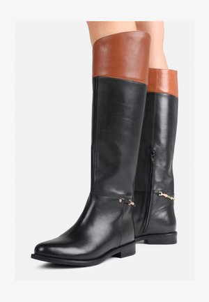 Bottes en cuir noires arrivant au genou avec des poignets bruns, des zips latéraux, des talons bas et de petites chaînes décoratives dorées, portées sur des jambes nues.