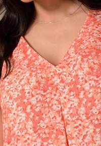 Blusa floreale corallo con scollatura a V, realizzata in tessuto leggero, con una superficie morbida e una combinazione di piccoli motivi floreali bianchi e rosa.
