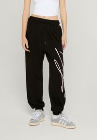 Svarta sweatpants med vida ben, elastisk midja och rosa grafisk tryck på sidan. Kombinerat med grå sneakers med vita detaljer.