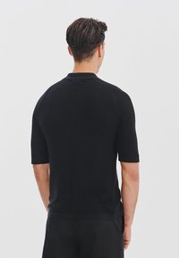 Mę�żczyzna z krótkimi ciemnymi włosami, ubrany w czarną koszulę z krótkim rękawem i czarne spodnie, stojący tyłem na jednolitym jasnym tle.