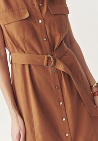 Vestido marrón de botones con mangas cortas, dos bolsillos en el pecho, un cinturón de tela con una hebilla de anillo dorado y una textura suave.