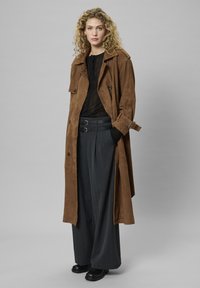 Göndör hajú nő, aki hosszú barna velúr esőkabátot visel fekete felsővel és szürke, bőszárú nadrággal, dupla csatos övvel és fekete csizmával.