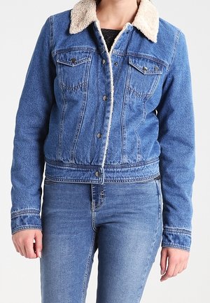 Kobieta ubrana w niebieską kurtkę jeansową z kremowym kołnierzem z polaru i niebieskie dżinsy, stojąca na tle jednolitego białego tła.