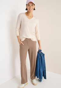 Beige V-Ausschnitt-Pullover, hellbraune Schlaghose, hält eine blaue Jeansjacke. Sneakers mit cremefarbenen und blauen Akzenten. Glatte Textur.