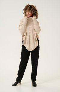 Beige Kabelstrick-Poncho mit Rollkragen, weitem Schnitt und strukturiertem Muster, kombiniert mit schwarzen schmalen Hosen und Stiefeletten mit Absatz.