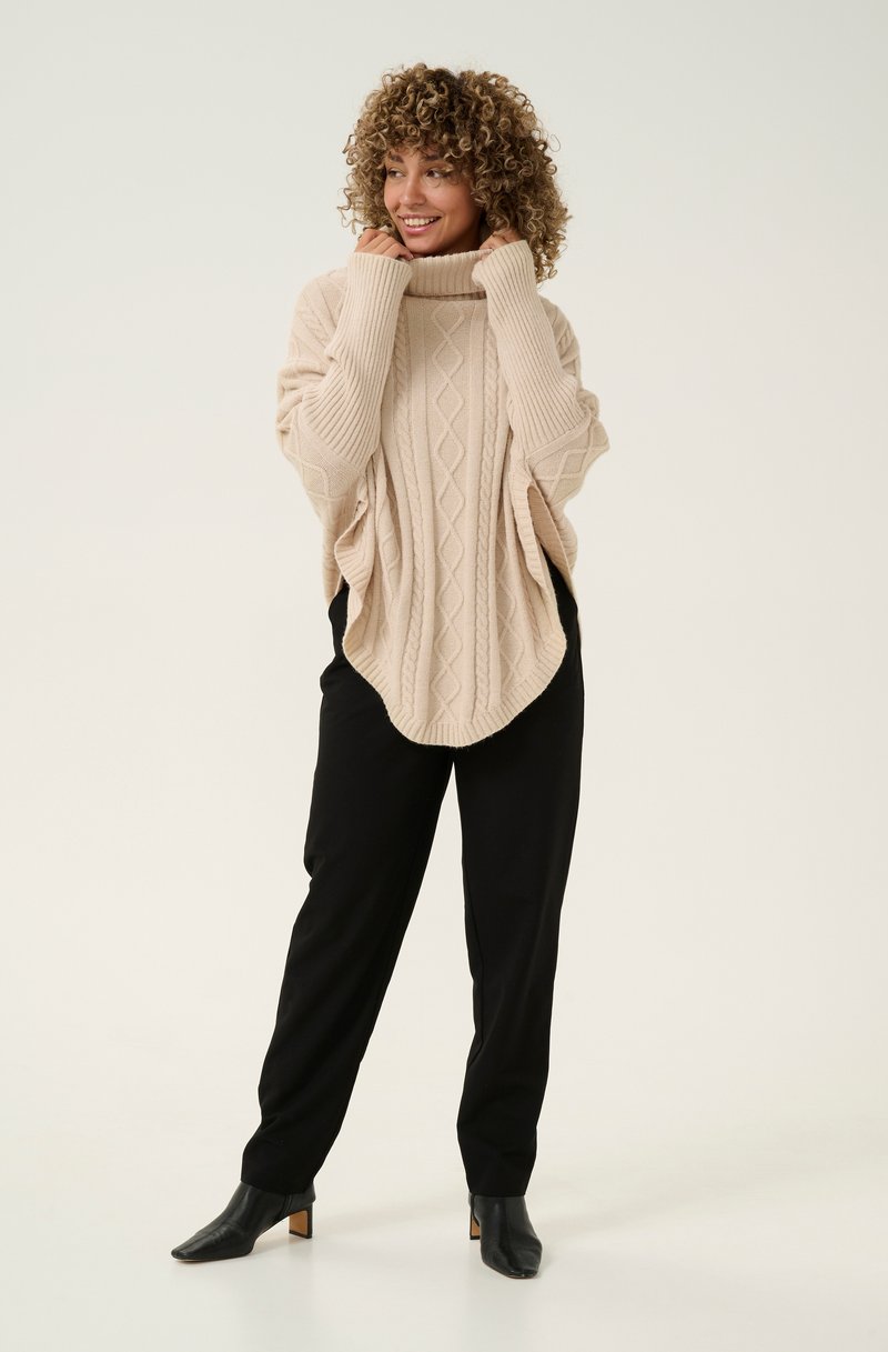 Beige Kabelstrick-Poncho mit Rollkragen, weitem Schnitt und strukturiertem Muster, kombiniert mit schwarzen schmalen Hosen und Stiefeletten mit Absatz.