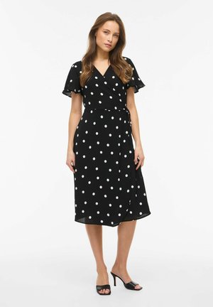 Kobieta w czarnej sukience kopertowej do kolan w białe kropki z falbaniastymi rękawami, w połączeniu z czarnymi sandałami na obcasie z odkrytymi palcami.