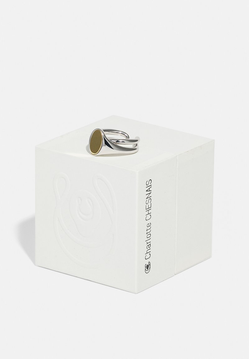 Ezüst állítható gyűrű arany ovális kiegészítővel, egy fehér, textúrázott dobozon bemutatva, amely domború mintázatú részletekkel rendelkezik.
