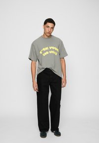 Šedé tričko s krátkým rukávem a žlutým grafickým textem, v kombinaci s černými širokými kalhotami a tmavými botami. Jednoduchý, neformální outfit.
