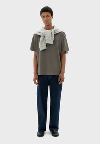 Grijs katoenen t-shirt met een ronde hals, gecombineerd met donkerblauwe denim jeans. Een lichtgrijze trui nonchalant over de schouders gedragen. Zwarte schoenen.