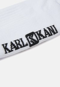 Chaussette blanche avec texture côtelée présentant un logo brodé noir "KARL KANI" sur le côté. Design simple et fonctionnel.