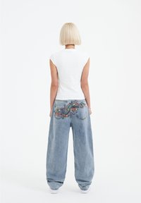White fitted t-shirt paired with loose, light blue denim jeans featuring colorful embroidered patterns on the back pockets.