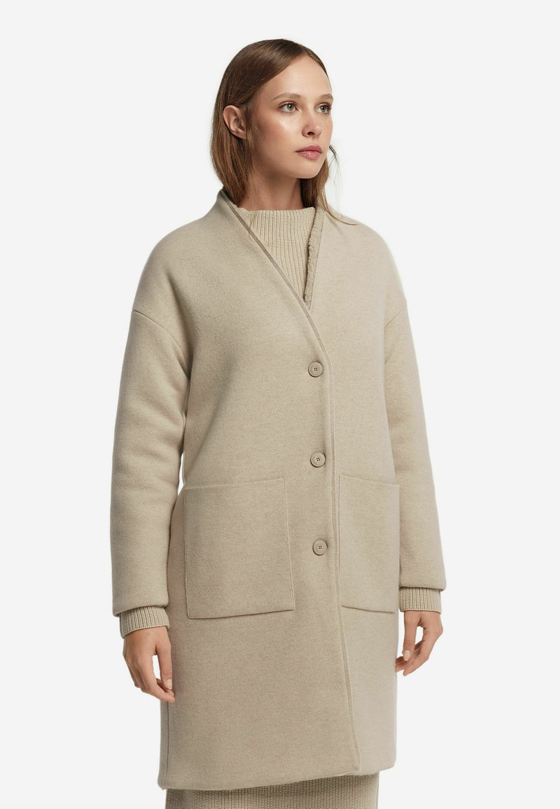 Cappotto lungo in misto lana beige con colletto a scialle, quattro bottoni e tasche frontali; presenta una texture morbida e un design caldo.