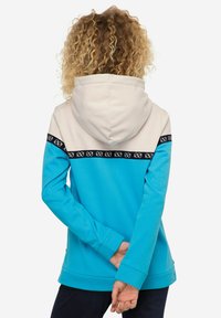 Sweat à capuche en turquoise et crème. Présente une bande noire avec un motif blanc sur la poitrine. Tissu doux. Manches longues.