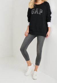 Donna che indossa un pullover nero GAP, jeans skinny grigi a vita alta e sneakers bianche, in piedi contro uno sfondo chiaro e uniforme.