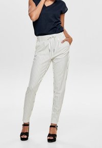 Femme portant un pantalon blanc à rayures verticales noires, un haut sans manches noir et des sandales à talons hauts noires, posant avec une main dans sa poche.