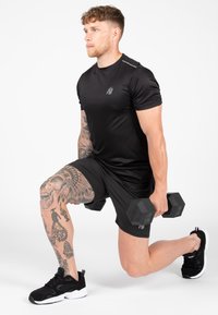 Juodas sportinis marškinėlis ir šortai, kuriuos dėvi raumeningas vyras, laikantis hantelius. Išsamios tatuiruotės ant jo kojos ir juodi sportiniai bateliai.