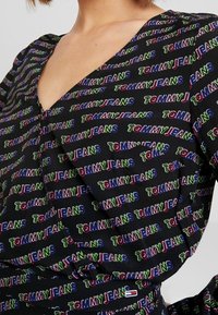 Blusa in tessuto nero con una grande stampa del logo "TOMMY JEANS" in rosso, verde e blu, con scollatura a V profonda e maniche corte.