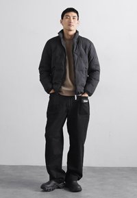 Casaco puffer preto com gola alta, usado sobre uma blusa bege. Combinado com calças soltas pretas e sapatos negros.