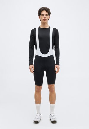 Fiatal férfi fekete, hosszú ujjú kerékpáros nadrágtartós rövidnadrágban, fehér pántokkal, fehér zokniban és fehér kerékpáros cipőben, egyszínű háttér előtt áll.