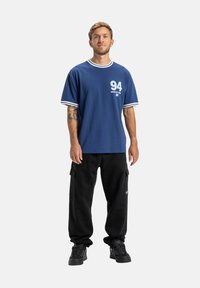 Námornícka modrá tričko s bielym lemovaním, so zobrazeným číslom "94" a logom. Vzhľad doplnený čiernymi cargo nohavicami a čiernymi teniskami s textúrovanou podrážkou.