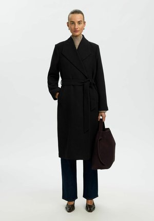 Femme portant un long manteau noir ceinturé sur un pantalon foncé, tenant un grand sac à main marron foncé, debout devant un fond clair uni.