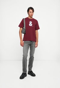Burgundijas krāsas kokvilnas T-krekls ar baltu ampersandu grafiku, apvienots ar pelēkiem džinsiem un melniem slip-on apaviem, papildināts ar mazu melnu somu.