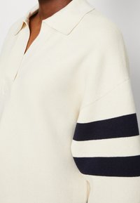 Zoom su una persona che indossa un maglione lavorato a maglia color crema con colletto e strisce orizzontali blu navy e crema sulle maniche.