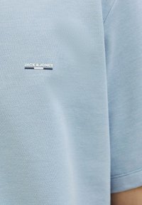 Svetlo modra polo majica iz teksturiranega materiala, z malim belim logotipom "JACK & JONES" na levi strani prsi.