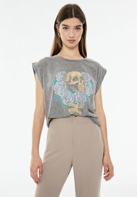 Camiseta sin mangas gris con un gráfico de calavera rodeado de rosas azules. Tela suave con un corte relajado y cuello redondo.