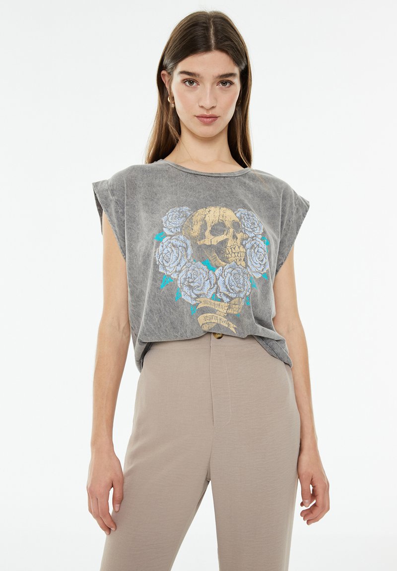 Camiseta sin mangas gris con un gráfico de calavera rodeado de rosas azules. Tela suave con un corte relajado y cuello redondo.