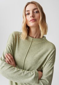 Blonde vrouw met blauwe ogen, gekleed in een lichtgroene langemouwtop, met gekruiste armen, tegen een effen lichtgrijze achtergrond.