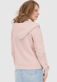 Felpa con cappuccio rosa di tessuto morbido, dotata di tasca a marsupio e polsini a costine, mostrata di spalle con jeans azzurri chiari.