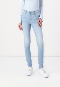 Jeans ajustados de color azul claro con zonas desgastadas, bolsillos delanteros y cierre de cremallera. Combinados con zapatillas blancas, sobre un fondo neutro.