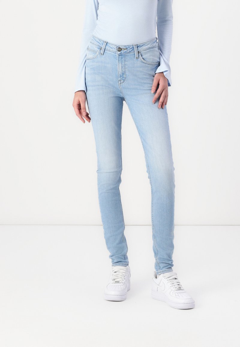 Jeans ajustados de color azul claro con zonas desgastadas, bolsillos delanteros y cierre de cremallera. Combinados con zapatillas blancas, sobre un fondo neutro.