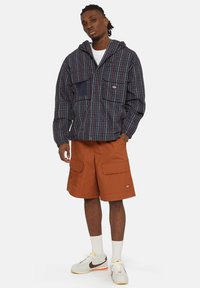 Giacca a quadri da uomo in blu navy e bordeaux con cappuccio, due tasche sul petto e polsini elasticizzati, abbinata a pantaloni cargo arancioni e sneakers.