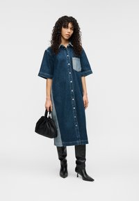 Τζιν φόρεμα με κοντά μανίκια, μπλοκ χρωμάτων σε ανοιχτό και σκούρο μπλε, κουμπιά μπροστά και τσεπάκι στο στήθος. Συνδυάζεται με μαύρη, ραμμένη τσάντα χειρός.