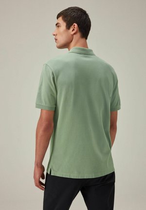 Polo verde de manga corta, hecho de una tela suave, con un cuello clásico, aberturas laterales y un ajuste relajado. Vista desde la parte de atrás.