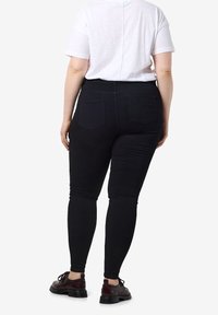 Personne debout, portant une chemise blanche à manches courtes, un jean skinny noir avec des poches arrière et des chaussures marron foncé à lacets.
