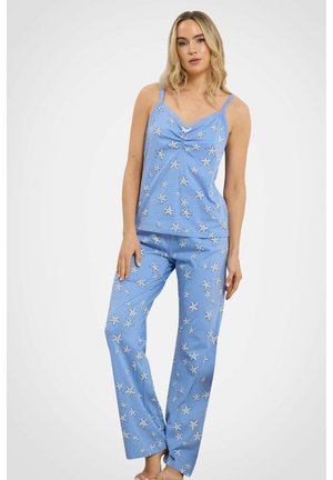 Donna che indossa un pigiama blu con motivo a stelle marine bianche, in piedi con una mano sulla coscia contro uno sfondo semplice.