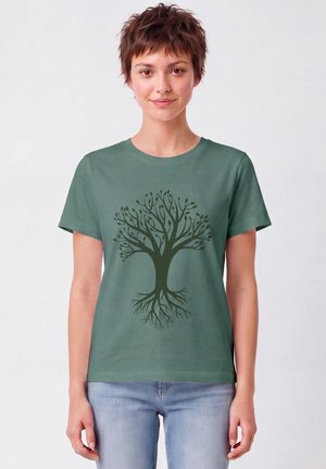 Junge Frau mit kurzen Haaren, die ein grünes T-Shirt mit dunklem Baum-Design und hellblaue Jeans trägt, steht vor einem einfarbigen Hintergrund.